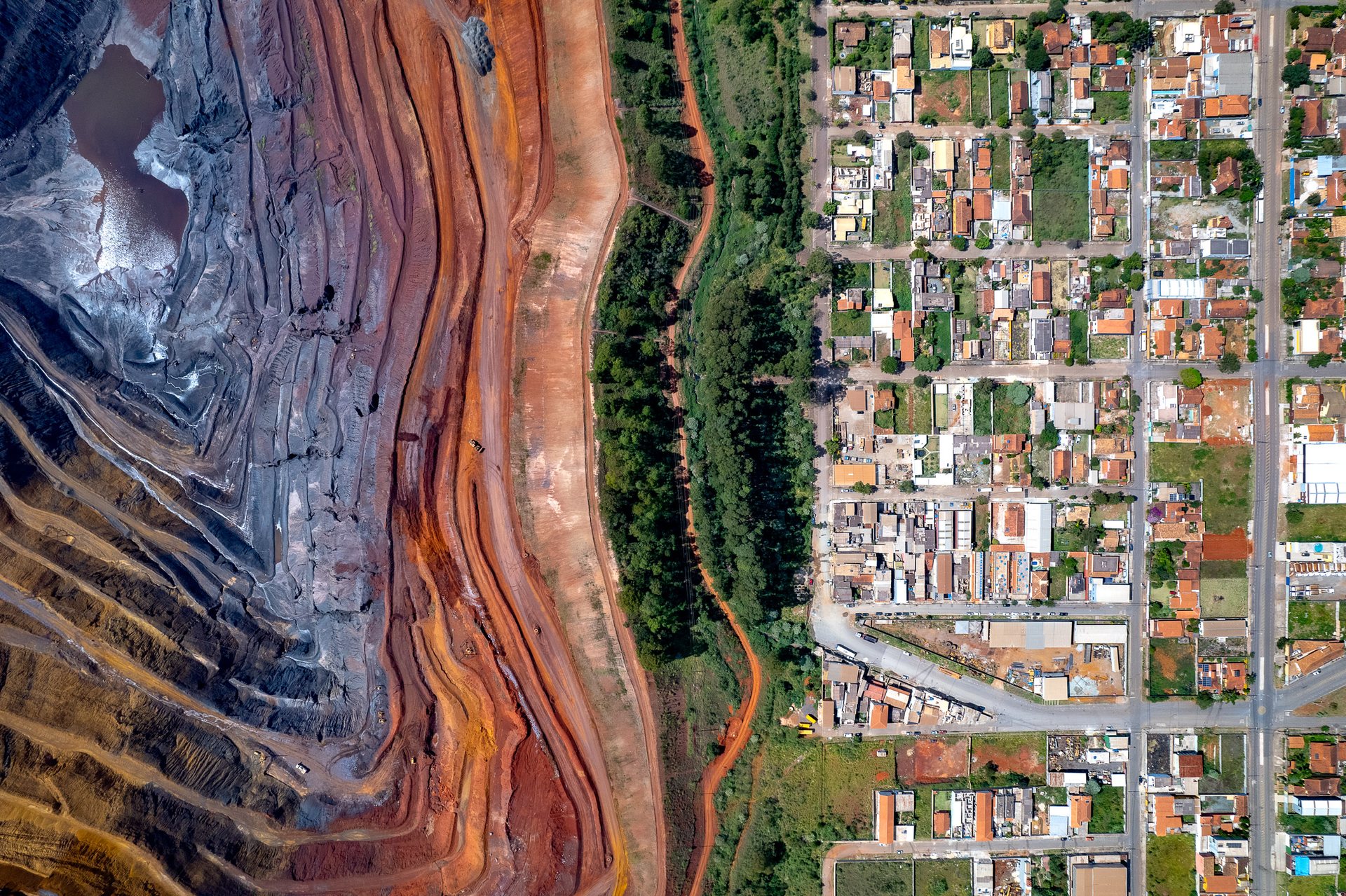 Aerial image showing a stark contrast between a massive open-pit mine with red and gray earth tones on the left, and a dense suburban neighborhood with houses and greenery on the right in Nova Lima, Minas Gerais.