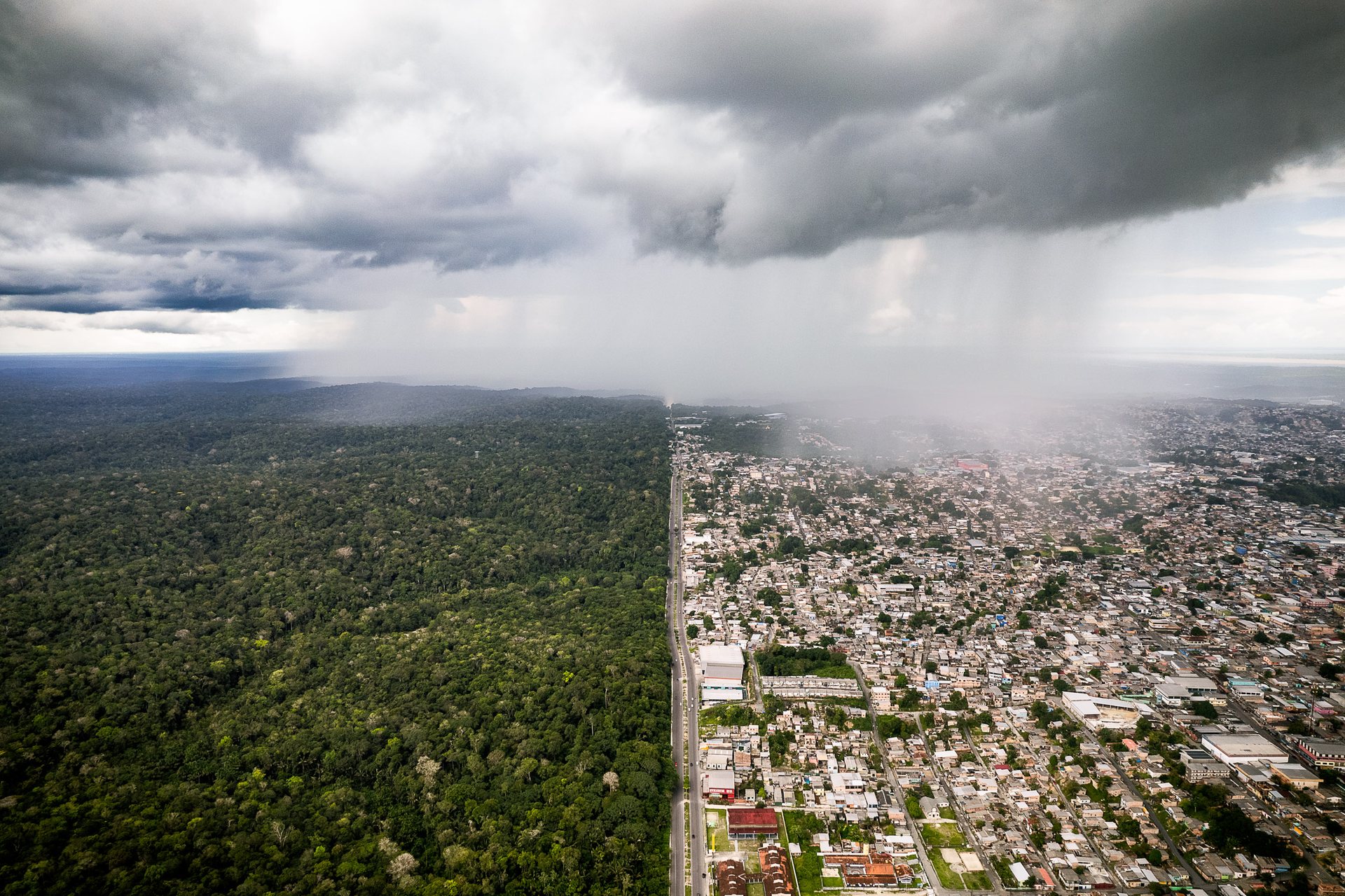 Aerial view of Manaus where a rainstorm moves across the boundary between the lush Amazon rainforest on the left and the urban cityscape on the right, highlighting deforestation.