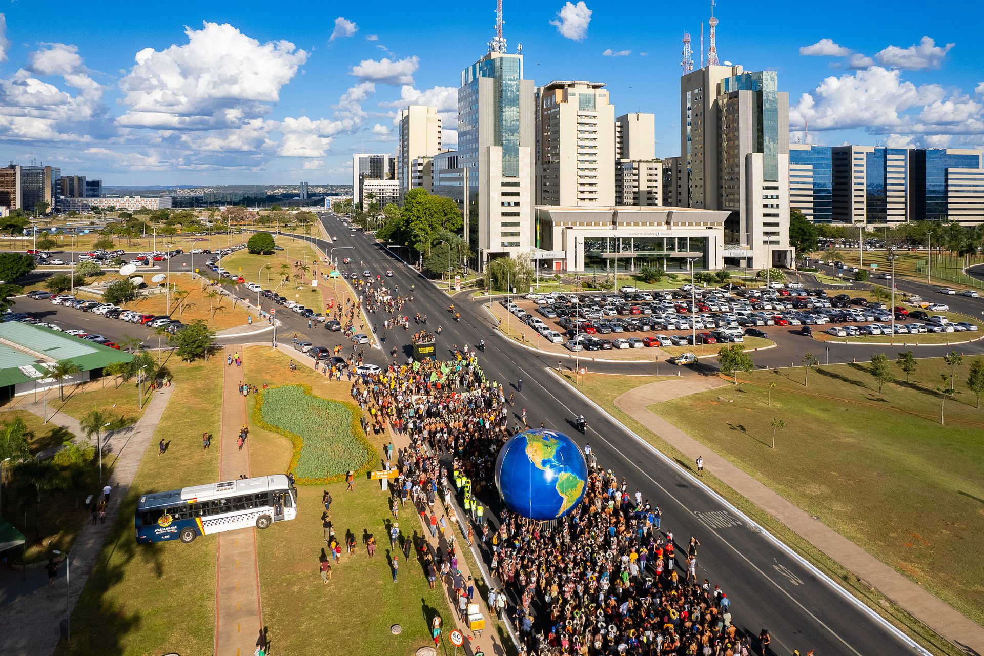 Thousands of Indigenous people march down a wide avenue in Brasília toward the National Congress during the Free Earth Camp protest, with government buildings in the background.