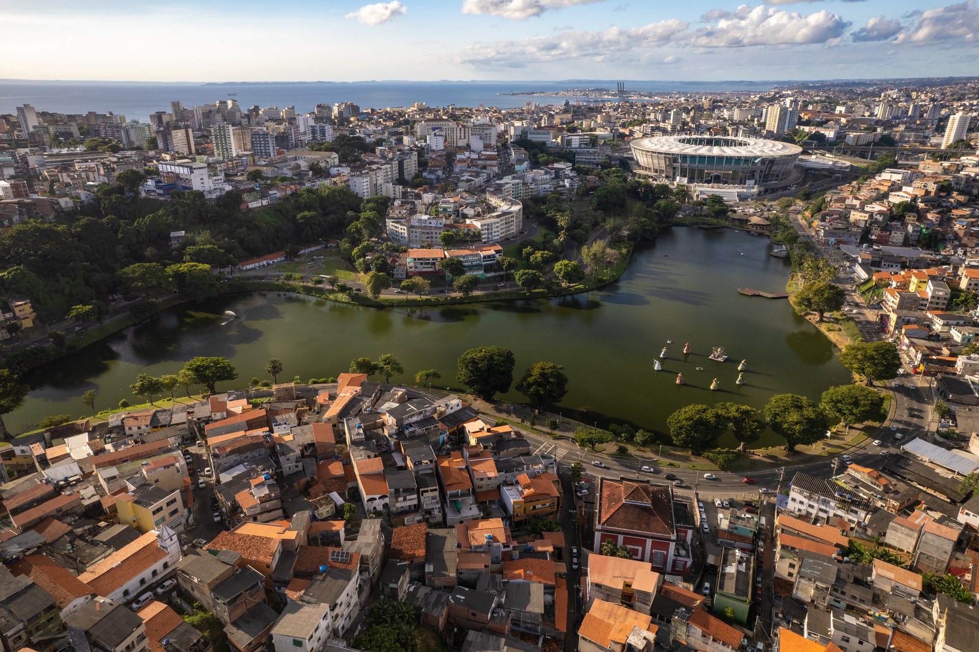 Aerial view of Salvador, Brazil, showing a dense favela foreground curving around Dique do Tororó lake with orixá statues in the water, backed by high-rise buildings and the Fonte Nova stadium in the distance under a bright sky.