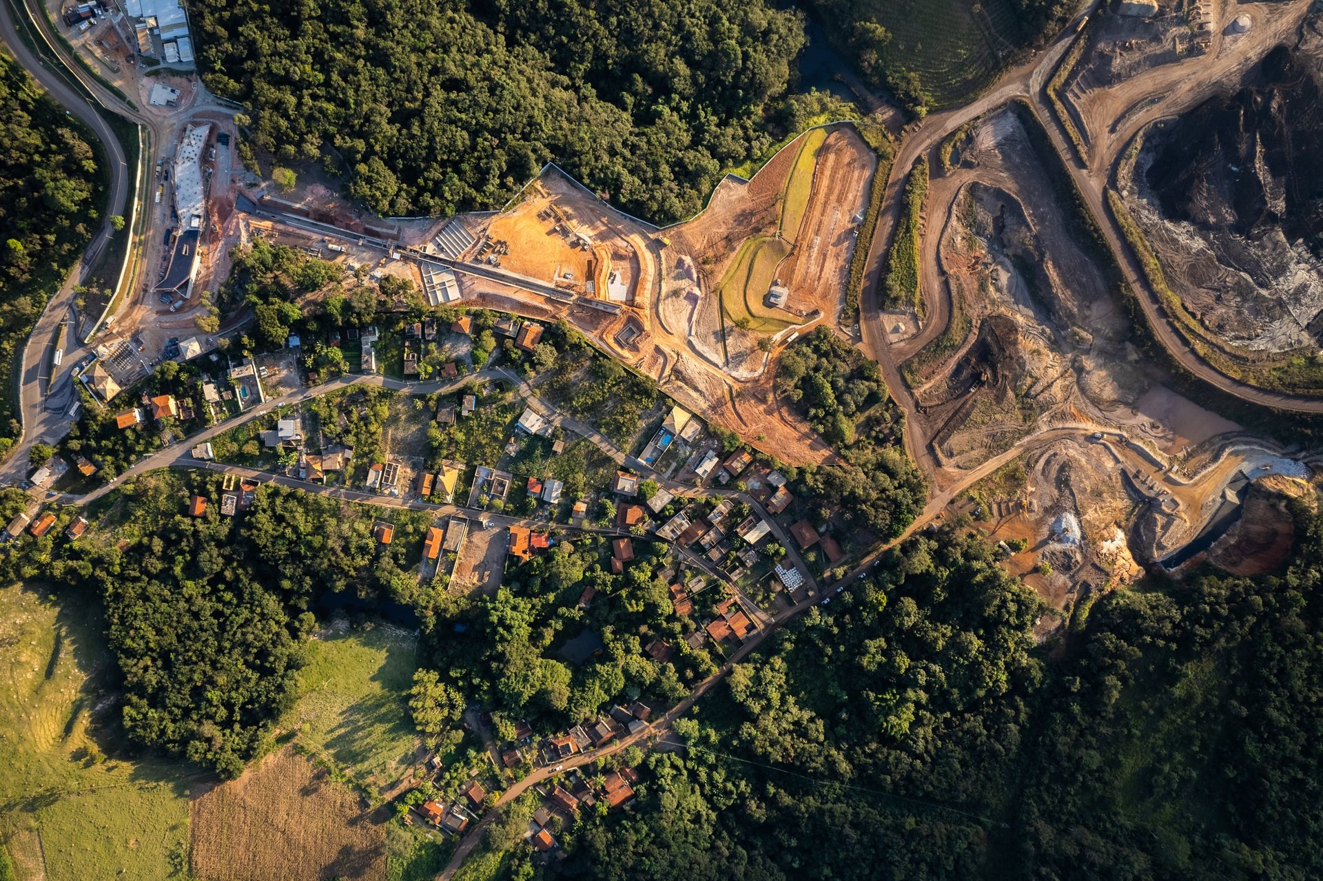 Aerial view of the Brumadinho region in Brazil where a mining dam collapsed in 2019, showing homes nestled beside extraction sites and lush forest, highlighting environmental and human impact.