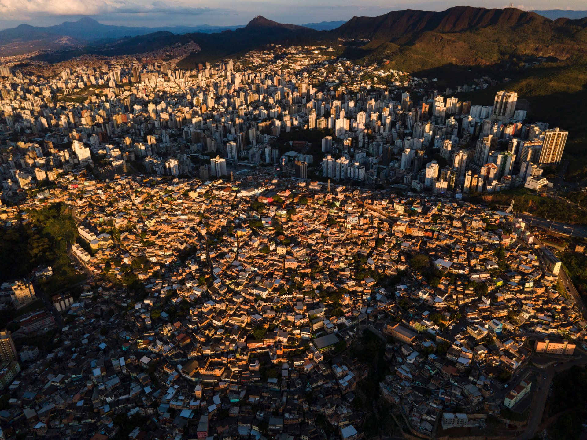 Dramatic sunset aerial of Morro do Papagaio favela in Belo Horizonte, Brazil, with golden light spilling over the hillside community as it merges into the formal cityscape beyond.