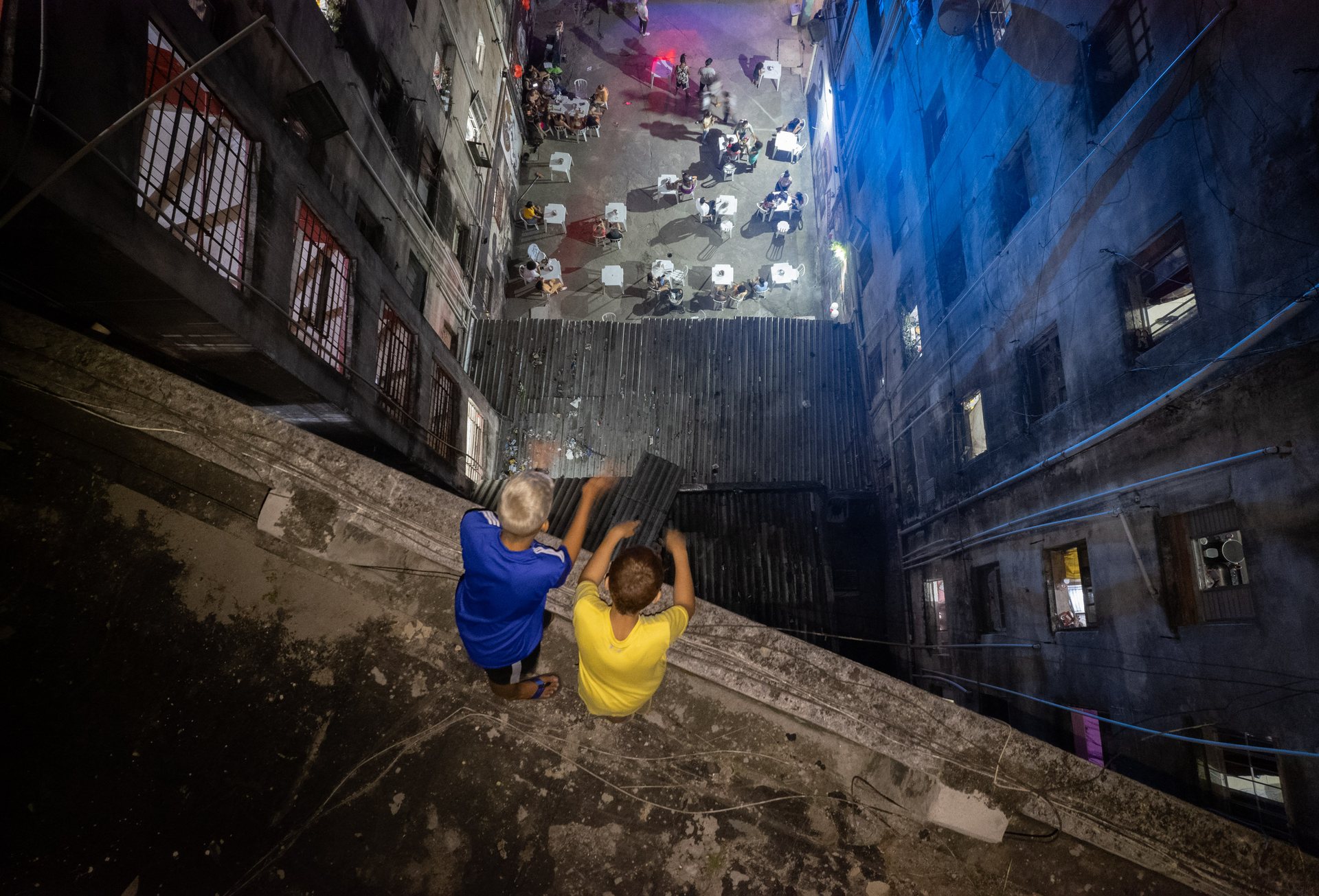 Downward view from the top of an occupied building in central São Paulo showing two men sitting on a ledge above a lit courtyard full of people celebrating below.