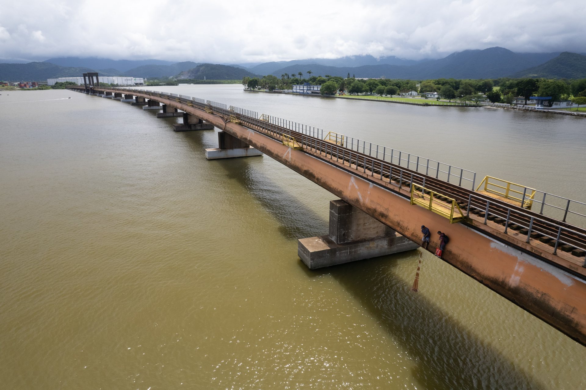 Oblique view of fishermen walking along a long, narrow rail bridge stretching across brown water in Santos, with mountains in the distance.