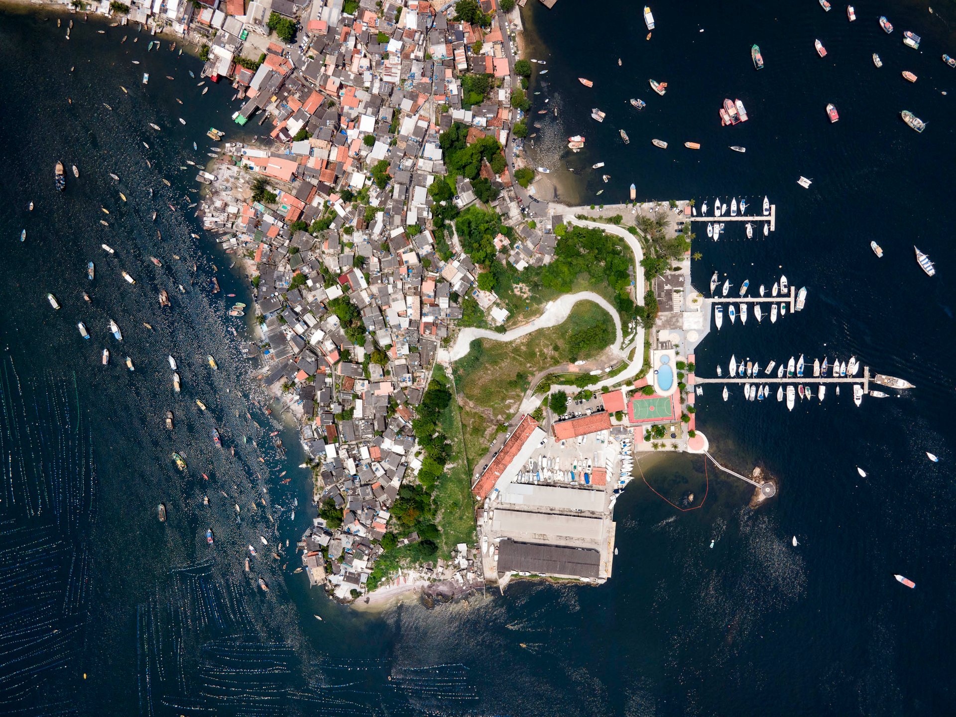 Top-down aerial showing a peninsula with the Jurujuba Yacht Club and an adjacent fishing colony across a narrow channel in Guanabara Bay, surrounded by boats.