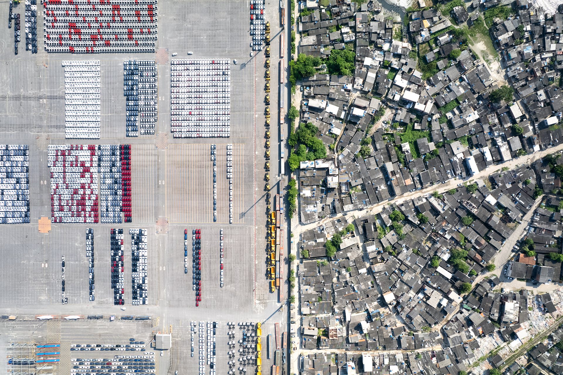 Direct aerial view of the Port of Santos showing tightly packed car lots on one side and a dense informal settlement on the other, split by a fence.