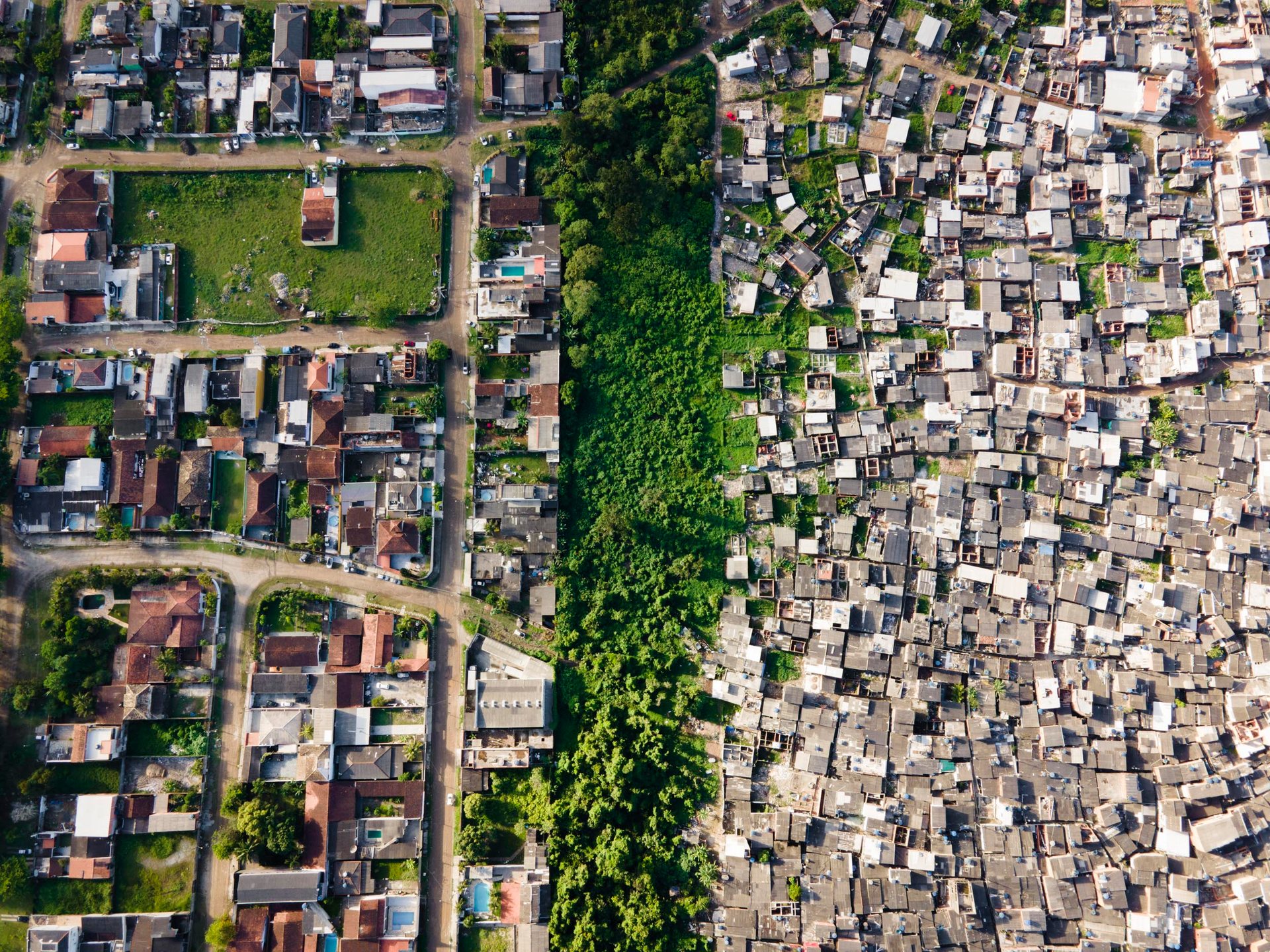 Clear aerial division between orderly, spacious formal housing with greenery and dense informal settlement, split by a thin strip of forest in Guarujá.