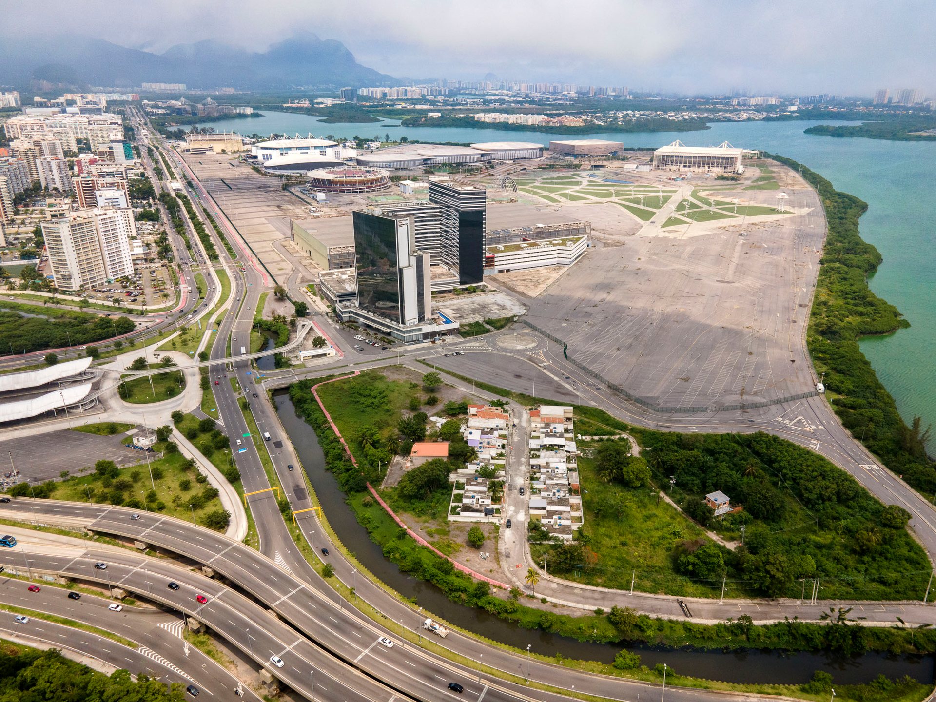 Wide aerial showing the remains of Vila Autódromo beside the sprawling 2016 Rio Olympics complex, with small homes surrounded by large-scale infrastructure and vacant land.