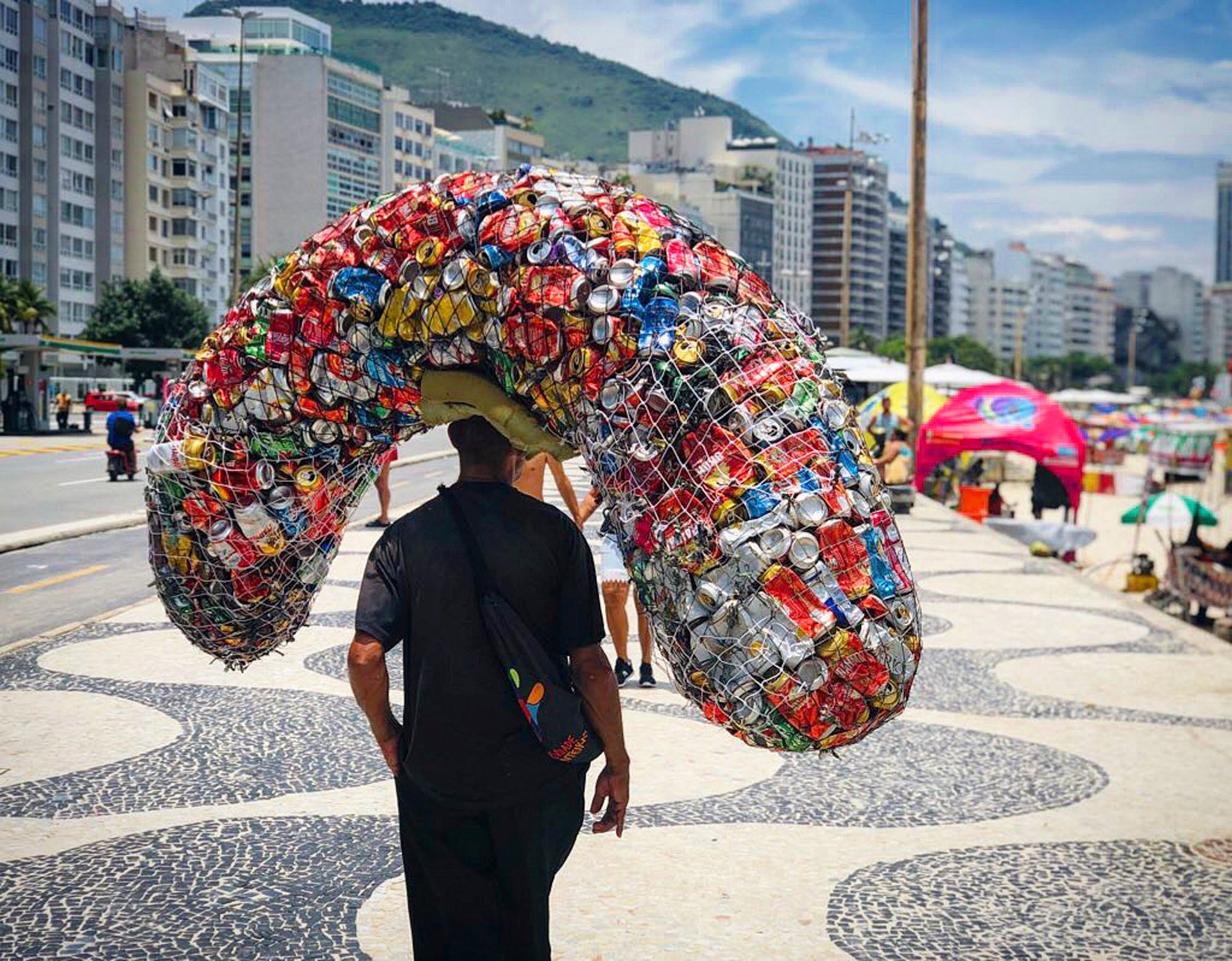 A man walks along Copacabana Beach in Rio de Janeiro carrying a massive load of crushed aluminum cans for recycling, framed by beachfront buildings and mosaic sidewalks.