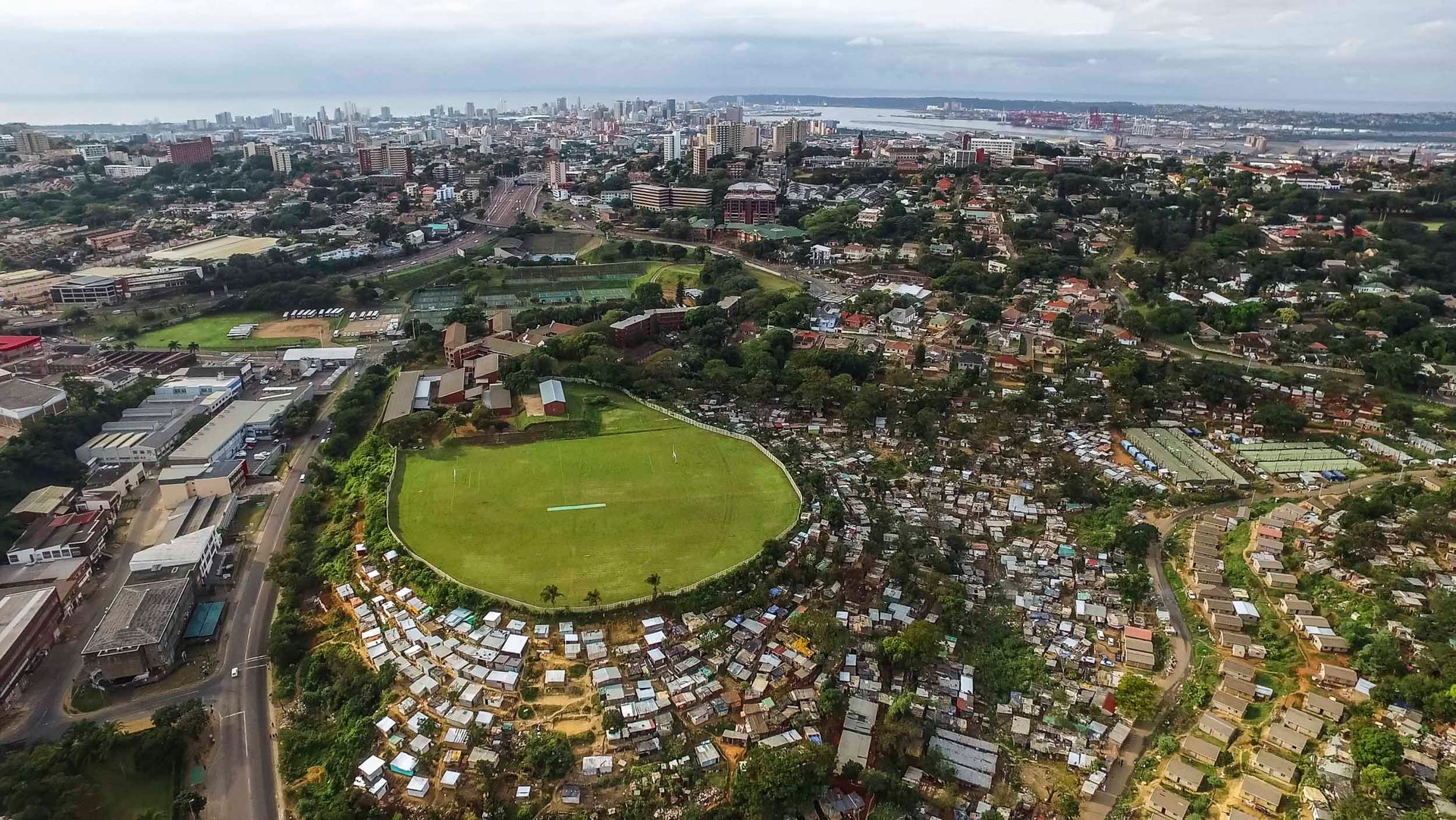 A cricket pitch surrounded by shacks, with the city center of Durban and the port in the background.