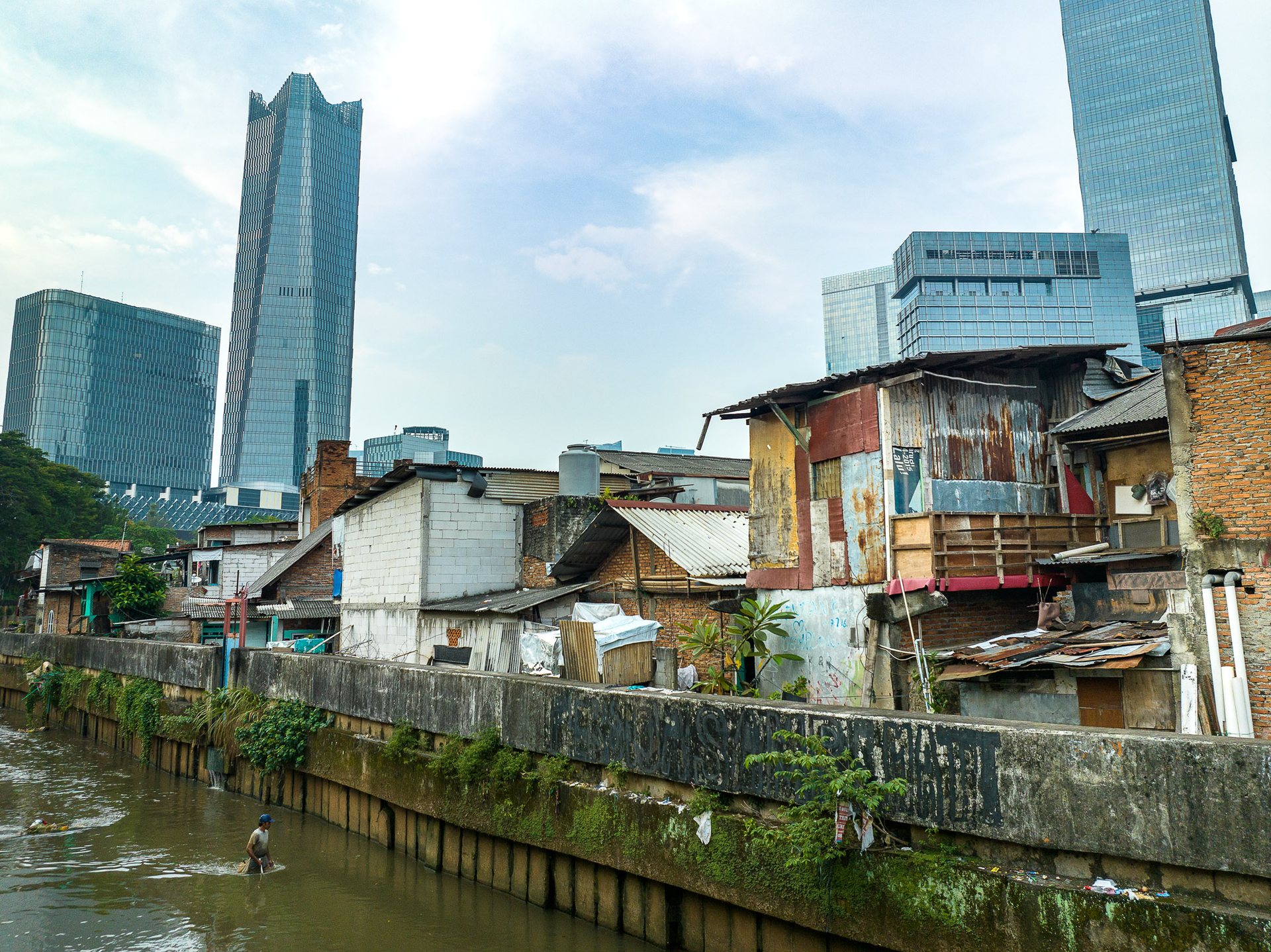 A man wades through a tributary of the Ciliwung River collecting plastic, in the shadow of high-rise buildings including the Four Seasons Hotel.
