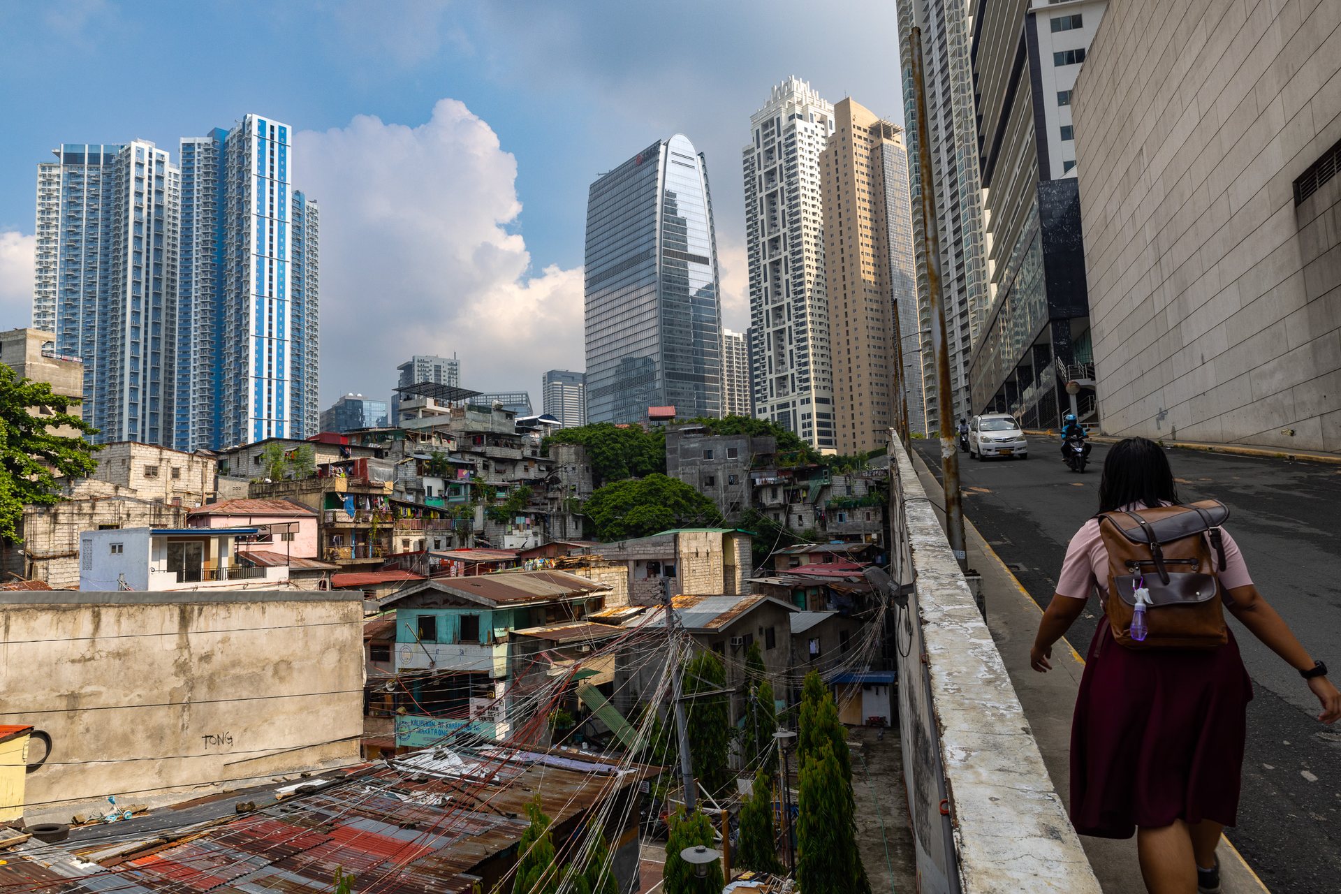 Contrasting cityscape in Manila with a woman walking through an alley bordered by high-rise towers of Bonifacio Global City and informal housing – capturing proximity of wealth and poverty.