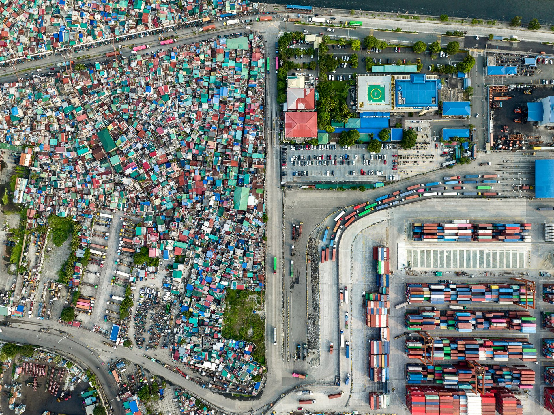 Top-down drone image of Manila showing a tightly packed informal neighborhood adjacent to a major shipping yard, bisected by a road filled with truck traffic entering the port.