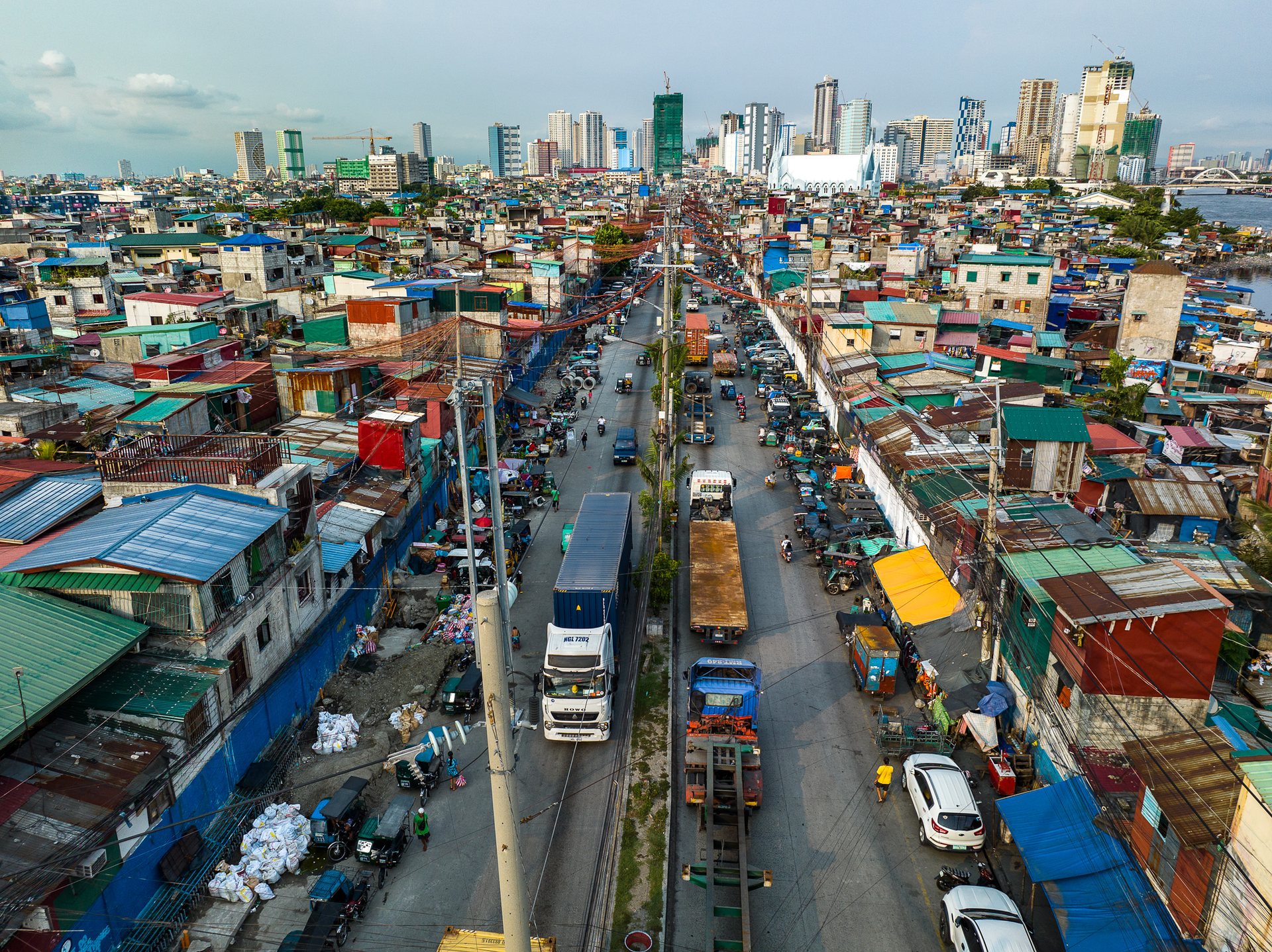 A busy road cutting through informal housing in Manila, crowded with trucks and power lines, leading toward the high-rise skyline in the background.