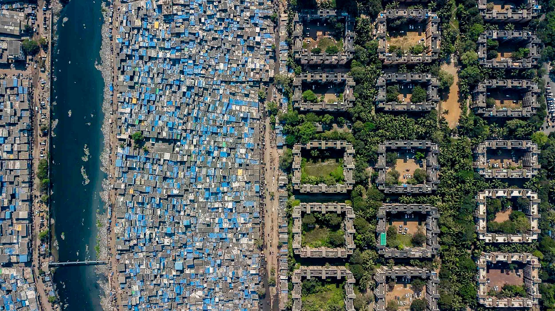 Aerial view of Mumbai showing dense blue-roofed slum adjacent to geometric luxury apartments near Bandra Kurla.