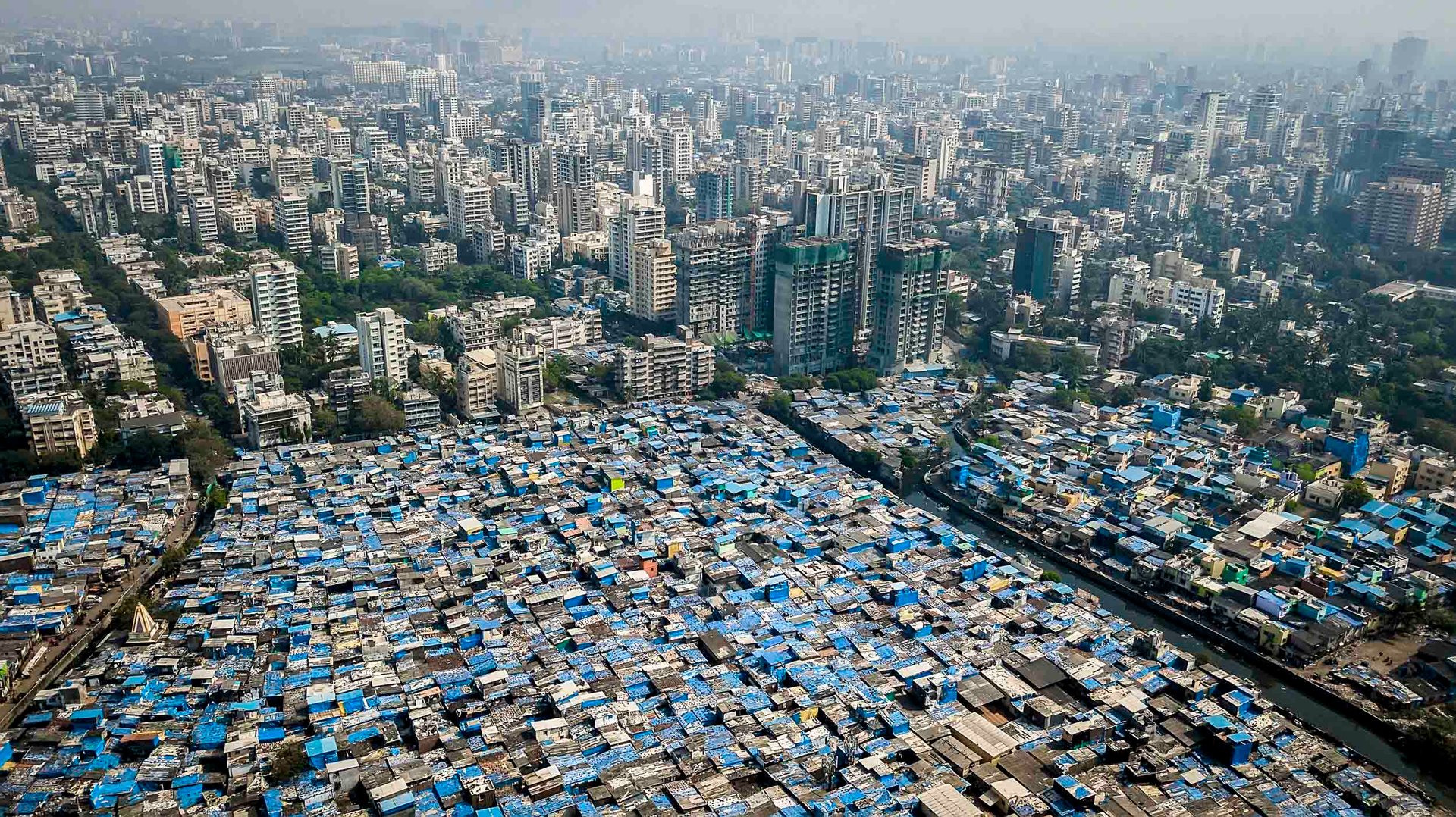 Slum settlement in Mumbai packed tightly against rising glass towers under haze.