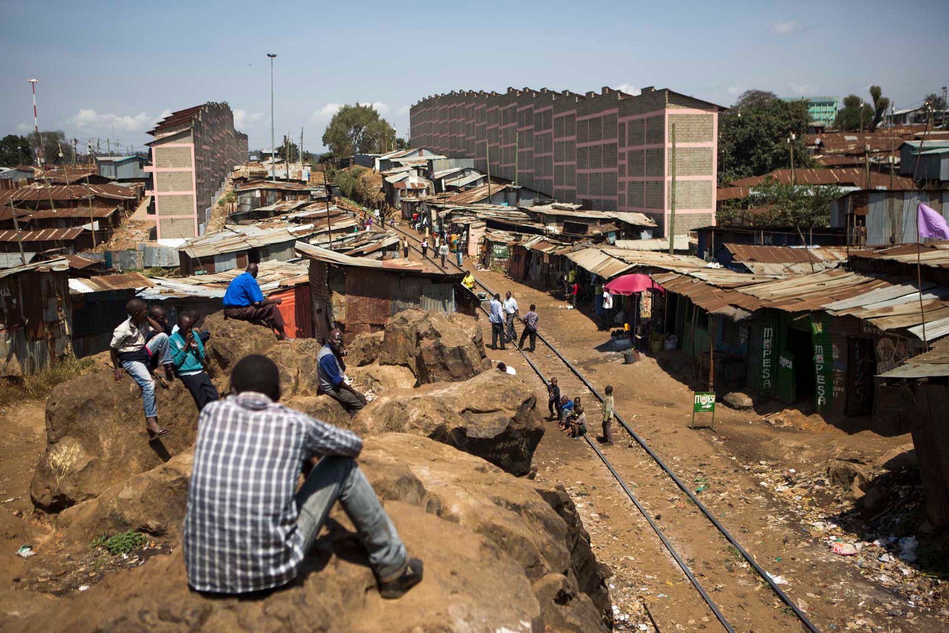 Photograph of Nairobi residents observing slum activity from a rocky outcrop above a busy settlement road.