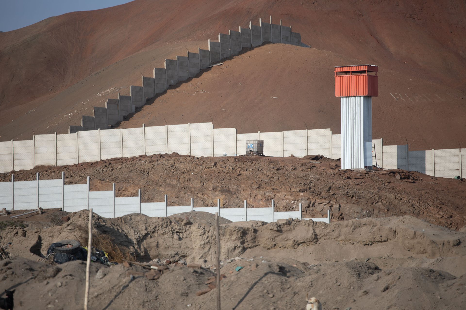 Concrete walls and a red guard tower cut through a barren desert hillside near Lima, Peru, marking boundaries even in remote areas.