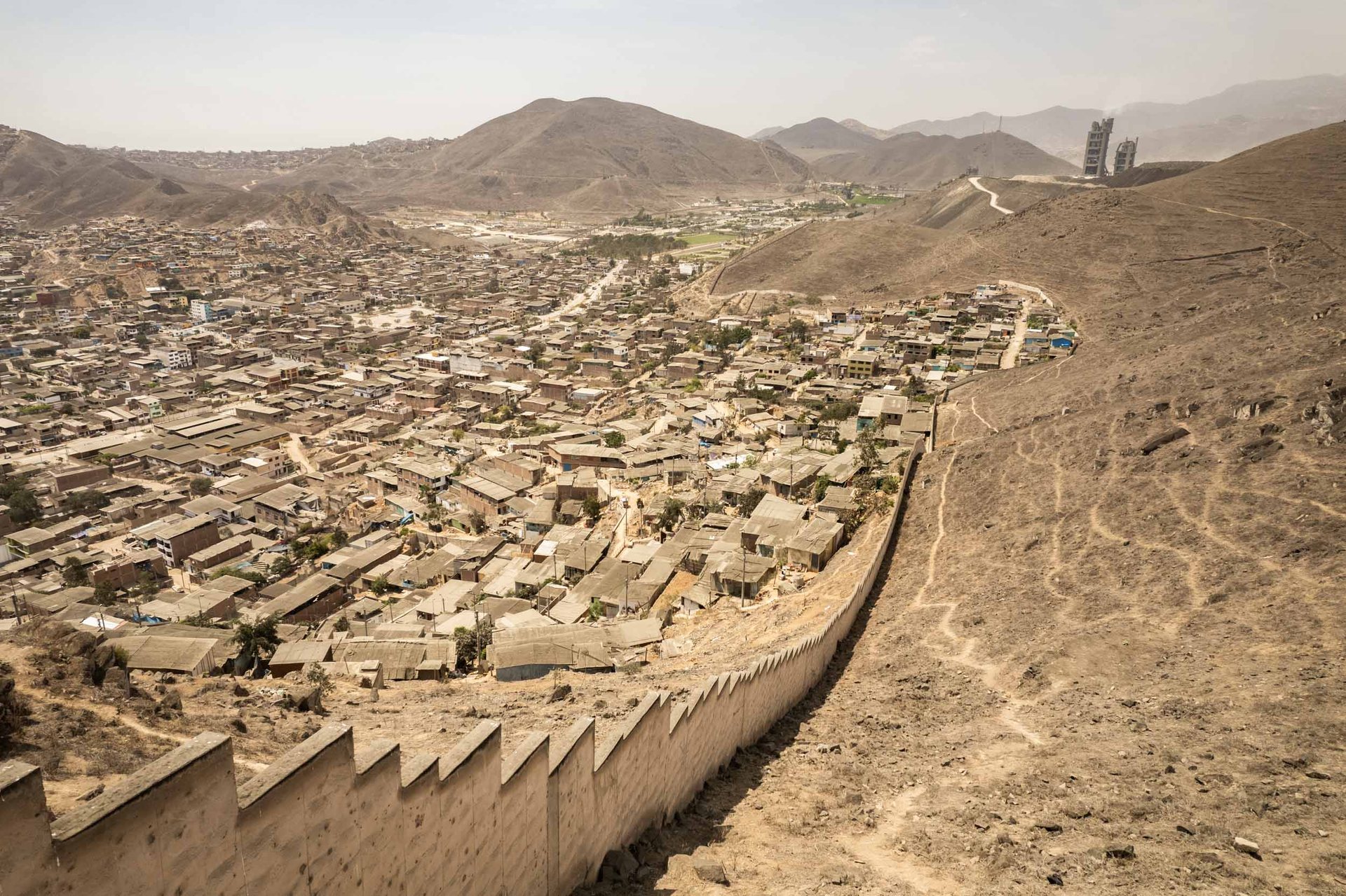 A walled division cuts through Villa Maria del Triunfo in Lima, separating densely packed homes from barren hills with a cement plant looming in the distance.