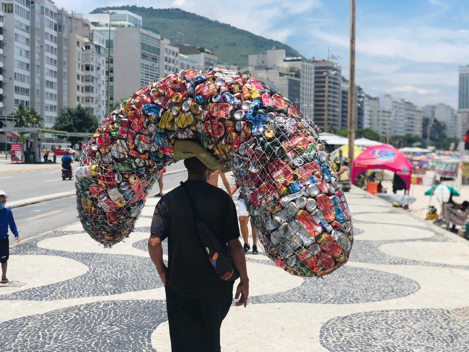 A man walks on Copacabana promenade with a large sack of aluminum cans on his head.