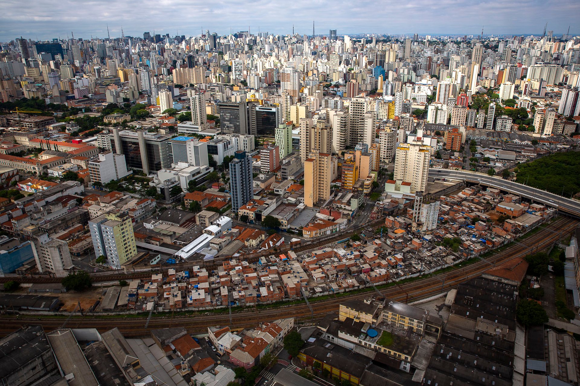 Panoramic aerial of São Paulo’s downtown core, with Moinho favela encircled by railway lines and towered over by a sprawling skyline of modern high-rise buildings, highlighting spatial inequality.