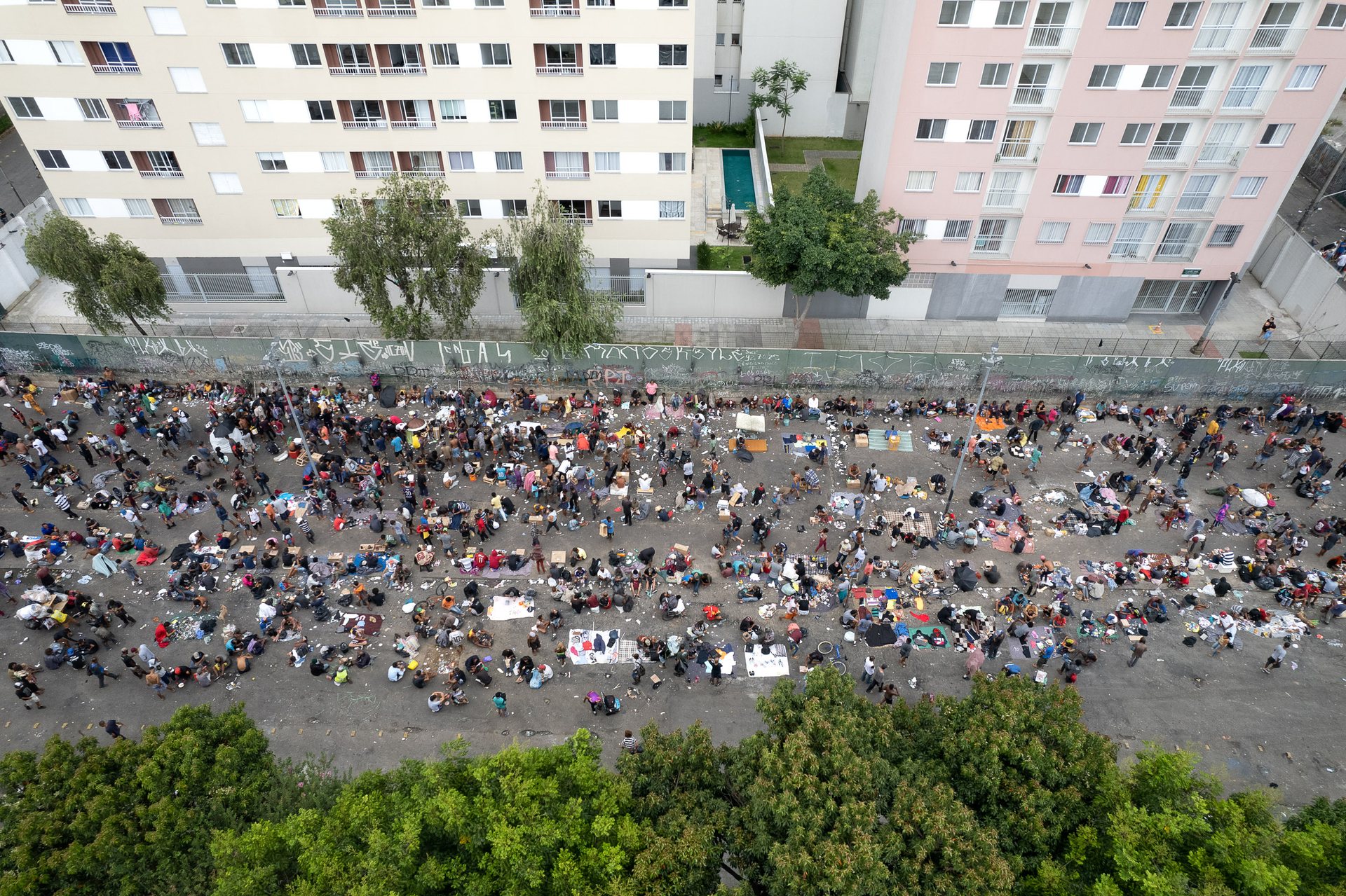 Overhead view of a large group of people gathered in São Paulo’s Cracolândia, an open-air drug-use area surrounded by public housing buildings and sparse greenery.