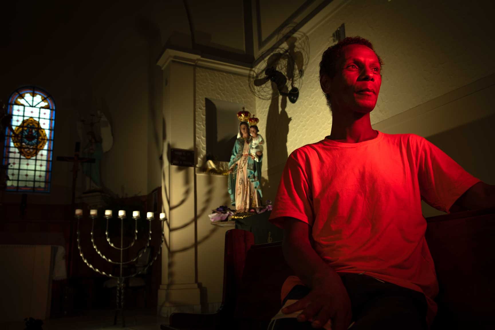 Portrait of Alessandra Martinelli, a trans woman living with HIV, sitting inside a dimly lit room in Mooca, São Paulo, with soft red light casting shadows on the wall behind her and a religious statue in the background.