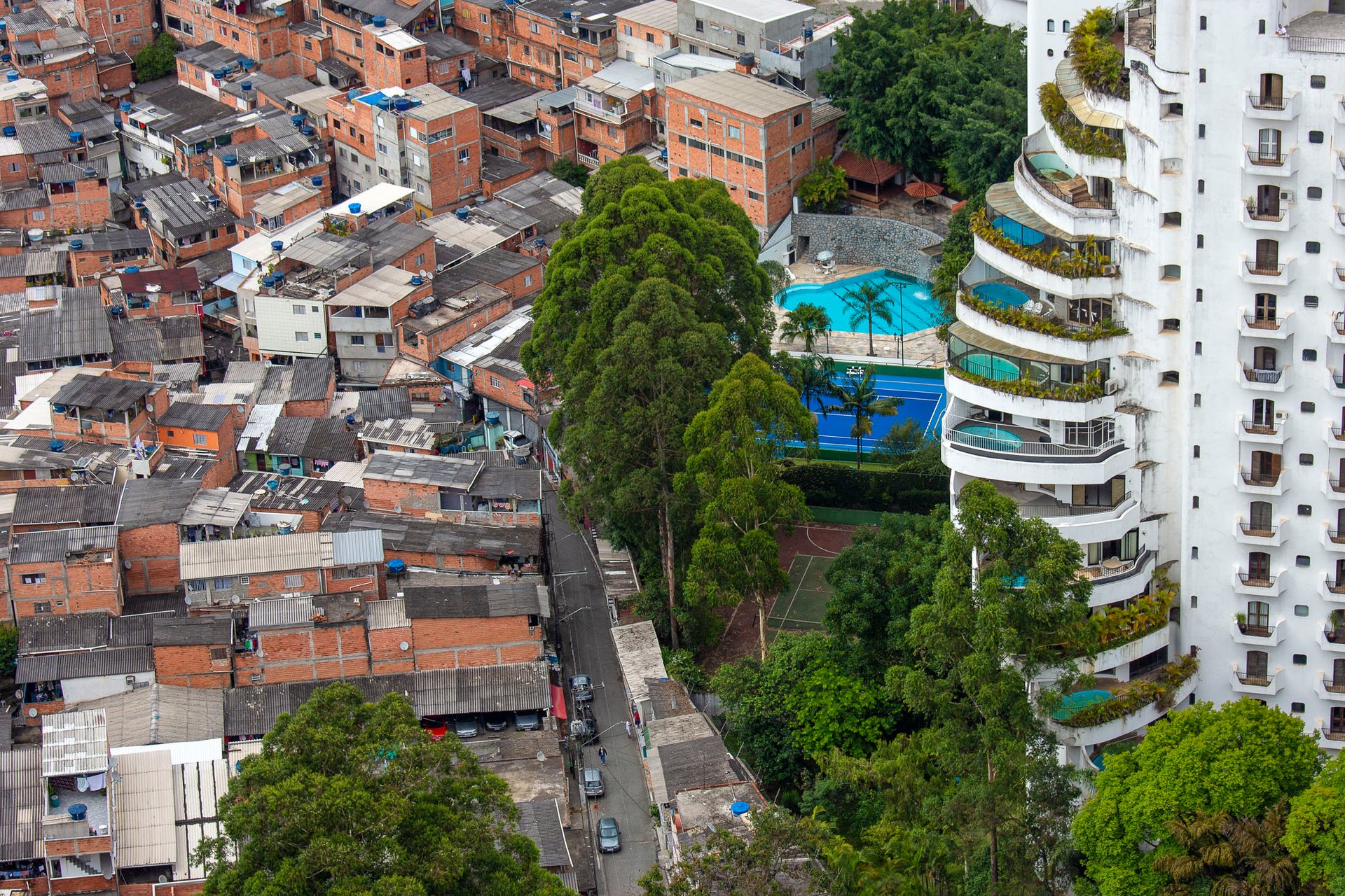 Aerial view of Paraísopolis favela in São Paulo, Brazil, showing dense red-brick homes directly beside a luxury high-rise tower with curved balconies and a private swimming pool, starkly illustrating urban inequality.