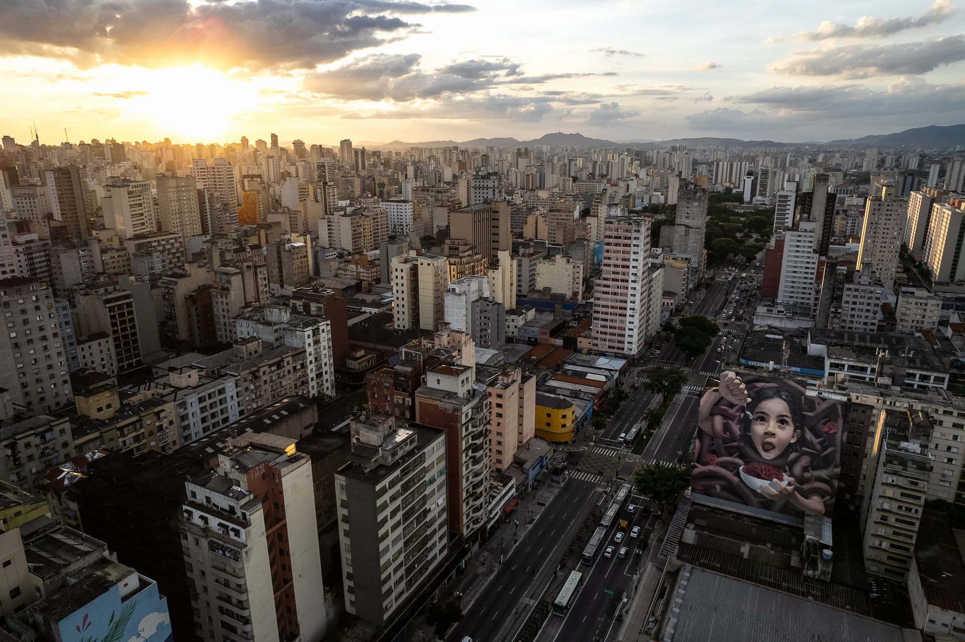 Wide cityscape of São Paulo at sunset, with a large mural of a girl eating cereal painted on a high-rise, juxtaposing innocence with urban sprawl.