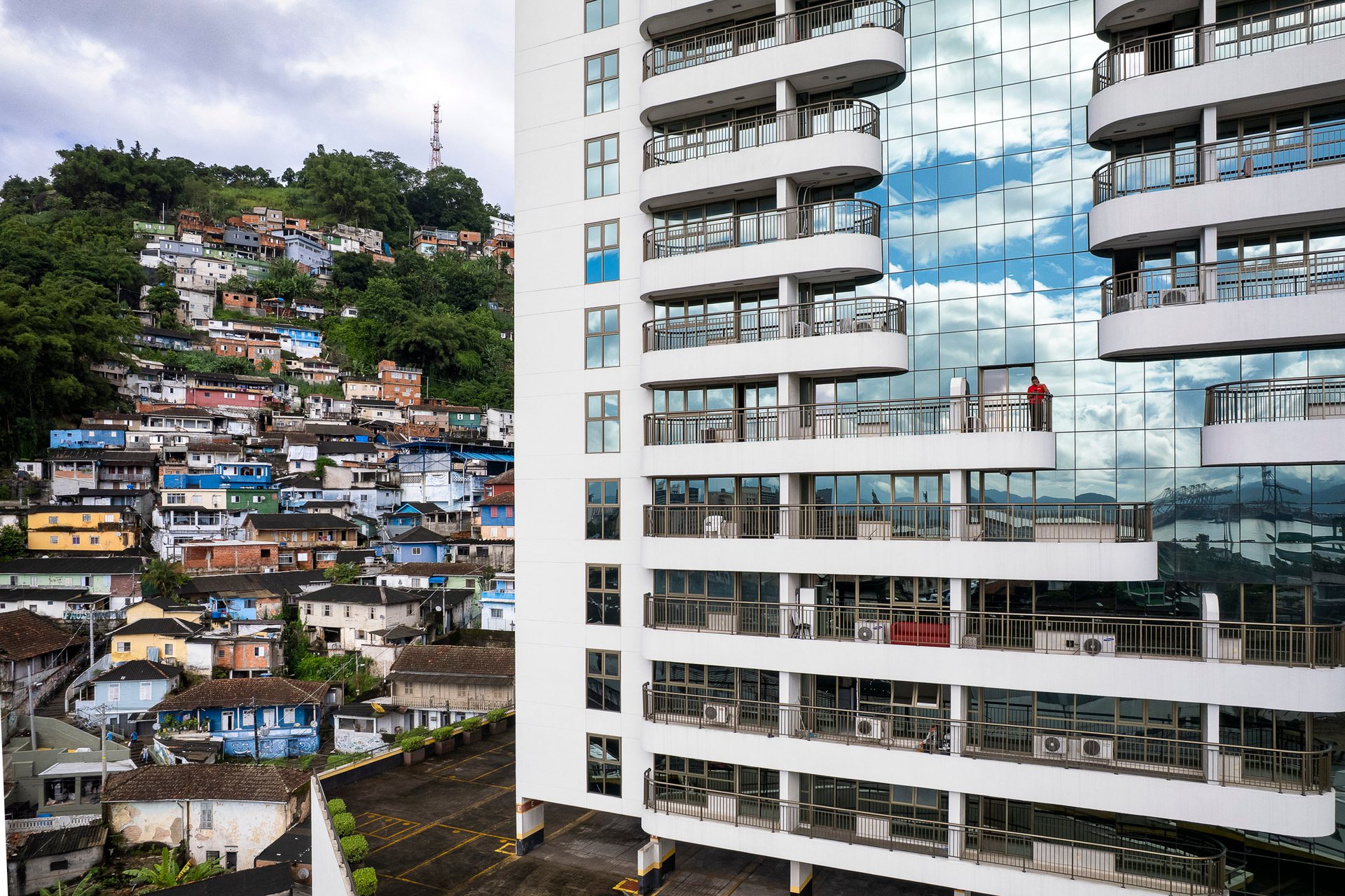 A stark visual of inequality as a favela clings to a forested hill while a mirrored high-rise reflects blue skies and private balconies in Santos.