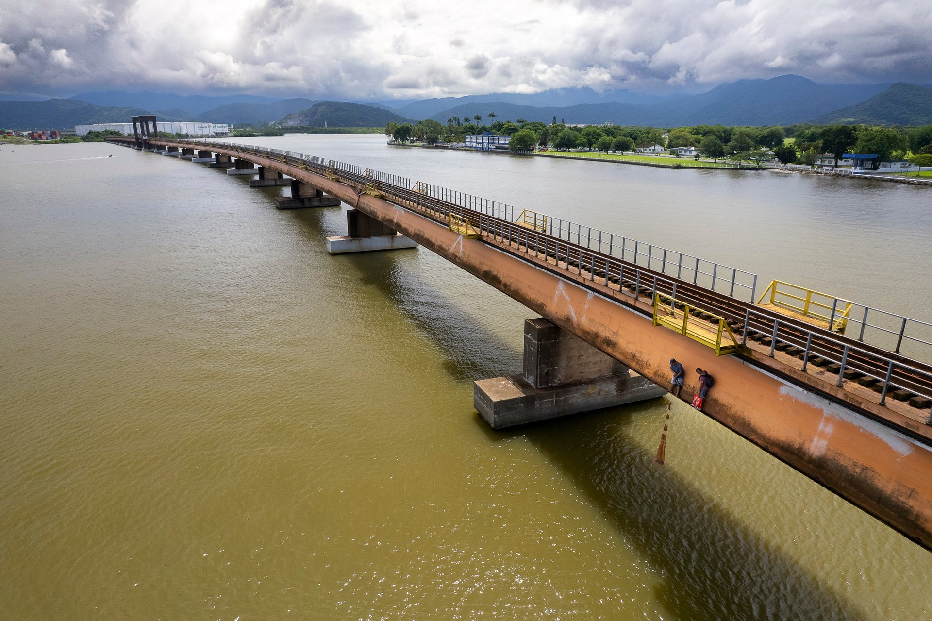 High above the estuary, fishermen walk along an old bridge in Santos, casting lines into muddy water as port development looms in the distance.